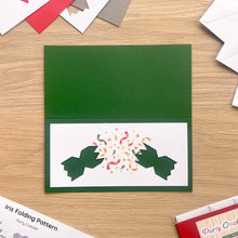 Load image into Gallery viewer, A green card blank with a white inner panel showing two cracker half cut-outs popping colourful paper confetti, photographed on a light wood tabletop with crafting papers around it.
