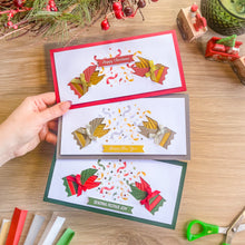 Load image into Gallery viewer, Three iris fold card designs are displayed on a wooden tabletop, held by a hand. The cards feature festive iris fold crackers in red, gold, and green, with confetti bursts and holiday greetings including 'Happy Christmas', 'Happy New Year', and 'Sending Festive Joy'
