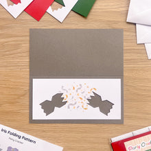 Load image into Gallery viewer, A party cracker aperture card shown on a wooden tabletop, with a grey card blank and a white front panel featuring two grey cracker shapes facing each other with confetti and stars in the centre. Partially visible surrounding cards and envelopes suggest a crafting setting.
