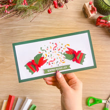Load image into Gallery viewer, A hand holds a handmade rectangular greeting card with a green border and festive iris fold cracker design with the text 'SENDING FESTIVE JOY'. The card is photographed on a light wood tabletop with Christmas decorations in the background.

