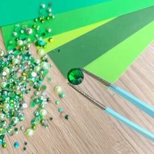 Load image into Gallery viewer, Green gemstone held by tweezers over a surface with green and wooden texture
