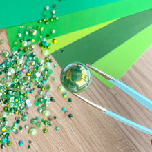 Load image into Gallery viewer, Green gemstone held by tweezers with green gem mix on a wooden surface
