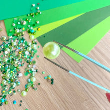 Load image into Gallery viewer, Green bead being held by tweezers with gem mix on a wooden surface
