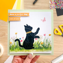 Load image into Gallery viewer, A handmade greeting card featuring a black iris fold cat silhouette in a grassy field with colorful flowers, a pink butterfly, and orange tabs that read 'Have a purrfect Day' and 'Lots of Love'. The card is held by a hand over a wooden craft workspace background.
