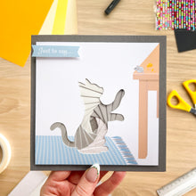 Load image into Gallery viewer, A handmade layered card featuring an iris fold cat on a blue-and-white checkered rug, held by a hand over a wooden desk with craft supplies.

