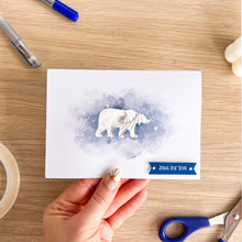 Load image into Gallery viewer, A handmade greeting card featuring a white iris fold polar bear on a blue watercolor background, held by a hand over a wooden desk with crafting supplies with 'Just for you' greeting.
