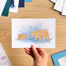 Load image into Gallery viewer, A hand holds a white art print featuring a watercolor blue background with two  polar bear cut-outs, shown on a light wood desk with other craft papers around it.
