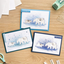 Load image into Gallery viewer, Three handmade iris fold greeting cards featuring watercolor polar bear illustrations are arranged on a light wood tabletop. The cards have different colored borders in blue, teal, and navy, and are surrounded by crafting tools like scissors, a stamp, and paper.
