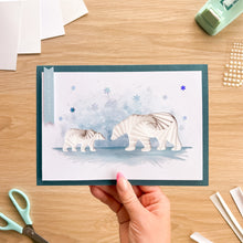 Load image into Gallery viewer, A handmade greeting card featuring two white polar bear silhouettes on a blue watercolor background, held by a hand over a wooden craft table with scissors and paper supplies.

