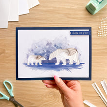 Load image into Gallery viewer, A handmade greeting card featuring a watercolor background and iris fold polar bears, held by a hand over a wooden desk with crafting tools.

