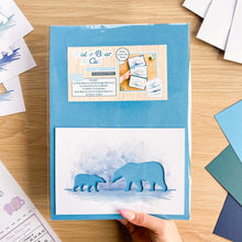 Load image into Gallery viewer, A hand holds a blue cardstock aperture pack on a wooden table, showing a sample polar bear cutout card inside. Scattered craft papers and cardstock sheets surround the workspace.

