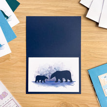 Load image into Gallery viewer, A navy blue greeting card featuring a watercolor-style polar bear illustration on a white lower panel, displayed on a light wood tabletop with other cards and stationery around it.
