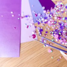 Load image into Gallery viewer, Close-up of a hand using tweezers to pick up a purple-pink gemstone on a wooden surface with scattered gems.
