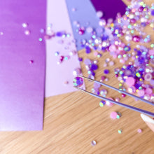 Load image into Gallery viewer, Colorful rhinestones being applied to a purple card with tweezers on a wooden surface.
