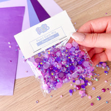Load image into Gallery viewer, Hand holding a bag of purple pearl gem mix with a wooden surface and purple paper in the background.
