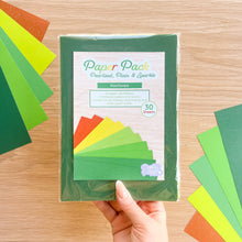 Load image into Gallery viewer, A hand holds a clear plastic-wrapped paper pack labeled 'Paper Pack Pearlised, Plain &amp; Sparkle' with 'Rainforest' as the color theme, surrounded by fanned sheets of green and bronze paper on a light wood surface.
