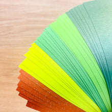 Load image into Gallery viewer, A close-up of a fanned stack of textured paper sheets in multiple shades of green and brown arranged on a light wood surface.
