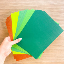Load image into Gallery viewer, A close-up of a fanned stack of textured paper sheets in multiple shades of green and brown arranged on a light wood surface.

