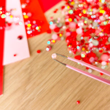 Load image into Gallery viewer, Pink tweezers with a red gem on a wooden surface with colorful blurred background
