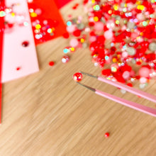 Load image into Gallery viewer, Close-up of red gems and a tool on a wooden surface with blurred colorful background

