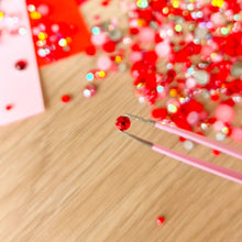 Load image into Gallery viewer, Close-up of red rhinestones and a tool on a wooden surface with blurred colorful background
