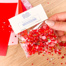 Load image into Gallery viewer, Hand holding a package of red pearl gem mix on a wooden surface with red paper in the background.
