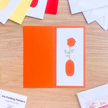 Load image into Gallery viewer, Card with a vase and rose design on an orange background, placed on a wooden surface.
