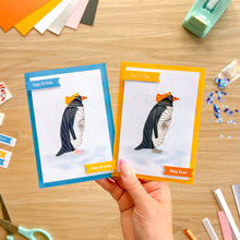 Load image into Gallery viewer, A hand holds two custom penguin-themed birthday cards over a wooden craft table. The left card has a blue border and says 'Happy Birthday' and 'Lots of Love', while the right card has an orange border and says 'Just To Say' and 'Stay Cool'.
