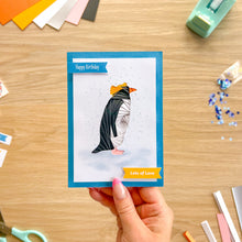 Load image into Gallery viewer, A hand holds a handmade iris fold greeting card featuring a penguin with orange head feathers. The card has a blue border and is displayed on a wooden desk surrounded by crafting supplies.

