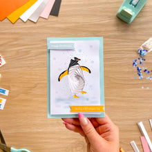 Load image into Gallery viewer, A handmade Christmas greeting card featuring an iris paper fold penguin, held by a hand over a wooden crafting desk with craft supplies.
