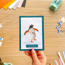 Load image into Gallery viewer, A handmade Christmas greeting card featuring an iris fold penguin, held by a hand over a wooden craft desk with colourful cardstock and crafting tools.
