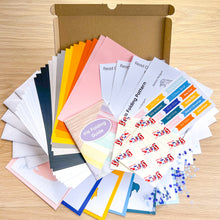 Load image into Gallery viewer, An open kraft cardboard box on a light wood surface contains a fanned-out set of card craft materials, including colorful paper sheets, reading guides, sticky notes, and a 'Iris Folding Guide'.
