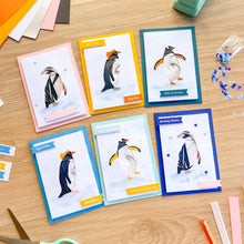 Load image into Gallery viewer, Six handmade greeting cards featuring penguin illustrations are arranged on a light wood desk with craft supplies like colored paper, scissors, and glitter nearby.
