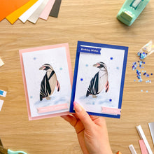 Load image into Gallery viewer, A hand holds two handmade greeting cards with penguin illustrations on a wooden table, surrounded by craft supplies like coloured paper, a tape dispenser, and snowflake confetti.

