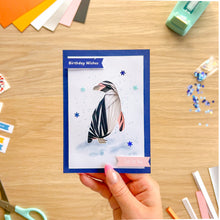 Load image into Gallery viewer, A hand holds a handmade greeting card featuring an iris fold penguin, set against a wooden desk with crafting supplies.
