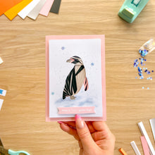 Load image into Gallery viewer, A handmade greeting card featuring an iris fold penguin, held by a hand over a wooden craft desk with colorful paper strips and stationery tools.
