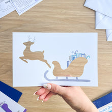Load image into Gallery viewer, Hand holding a card aperture with reindeer and sleigh design on a wooden surface.
