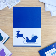 Load image into Gallery viewer, Blue and white card with reindeer and sleigh design on a wooden surface
