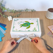 Load image into Gallery viewer, Person opening a white box with a turtle design on a wooden table.
