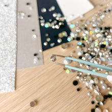 Load image into Gallery viewer, Close-up of a wooden surface with tweezers and scattered rhinestones.
