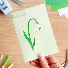 Load image into Gallery viewer, Hand holding a card with a snowdrop illustration on a wooden surface with stationery items.
