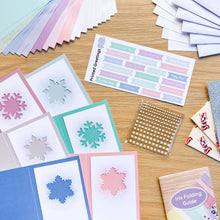 Load image into Gallery viewer, A flat lay of pastel greeting cards, paper cutouts, sticker sheets, and folding guides arranged on a light wood tabletop. Visible items include snowflake-cut cards, a sheet labeled 'Printed Greetings' with pastel labels, a 'Iris Folding Guide,' and stacks of folded paper.

