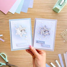 Load image into Gallery viewer, A hand holds two layered paper greeting cards with geometric snowflake designs and 'Happy Christmas' and 'Lots of Love' tags, set against a wooden crafting table with scissors, paper strips, and a tape dispenser.
