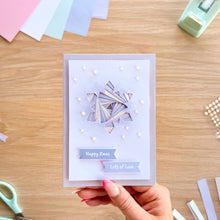 Load image into Gallery viewer, A handmade greeting card with a geometric layered snowflake design in lavender, white, and gold, held by a hand over a wooden desk with crafting supplies.
