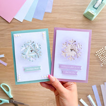 Load image into Gallery viewer, A hand holds two handmade greeting cards with snowflake designs on a wooden crafting table. One card has a mint green border and the other has a lavender border, both with white centers and decorative text banners.
