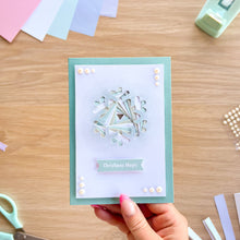 Load image into Gallery viewer, A handmade Christmas greeting card held by a hand, featuring a white snowflake design with pearl accents, a mint green border, and a banner reading 'Christmas Magic'. The card is photographed on a wooden desk with crafting supplies like scissors, pearl beads, and colored paper strips visible around it.
