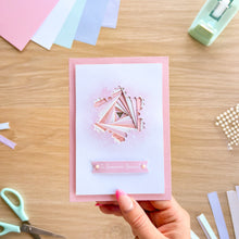 Load image into Gallery viewer, A handmade layered paper card with a pink and white design, held by a hand over a wooden desk with crafting supplies.
