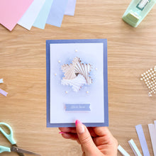 Load image into Gallery viewer, A handmade greeting card held by a hand, featuring an iris fold snowflake on a light blue watercolor background with the text 'Let It Snow'. The card is set against a wooden desk with crafting supplies like scissors, paper strips, and a tape dispenser.
