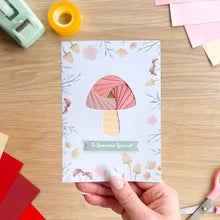 Load image into Gallery viewer, Hand holding a card with a mushroom design on a wooden surface with craft supplies.
