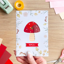 Load image into Gallery viewer, Handmade card with red mushroom design held by a hand on a wooden table with crafting supplies.
